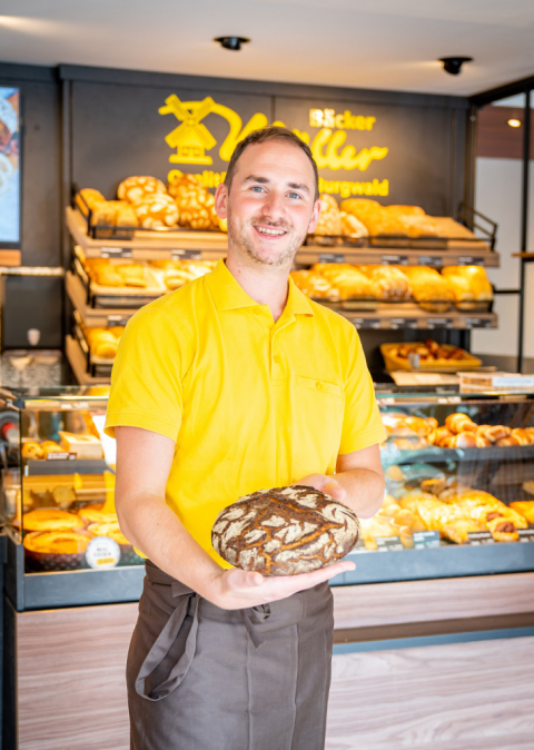 Bäcker Müller, Bild Verkaufstheke, der Verkäufer hält ein Brot in der Hand