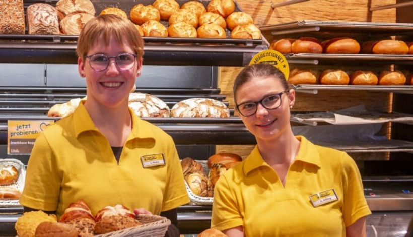 Bäcker Müller Azubi-Tag, Produktherstellung: zwei junge Frauen, die eine mit einem Brötchen und Snacks gefüllten Korb in der Hand, die andere mit einem Backblech, auf dem Backblech sind frische Brötchen